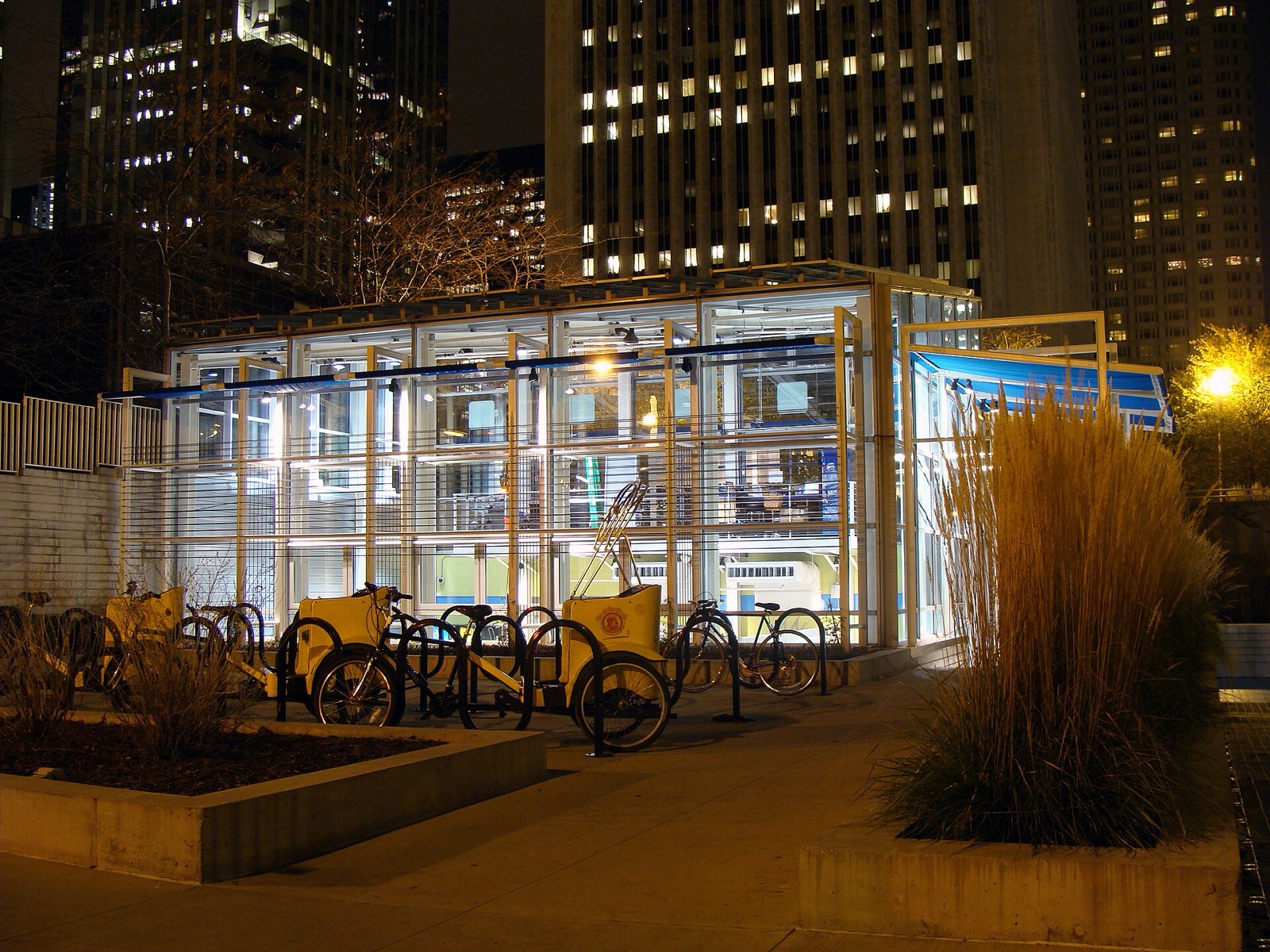 Rows of bike racks on the plaza in front of the lit glass bike station building at night